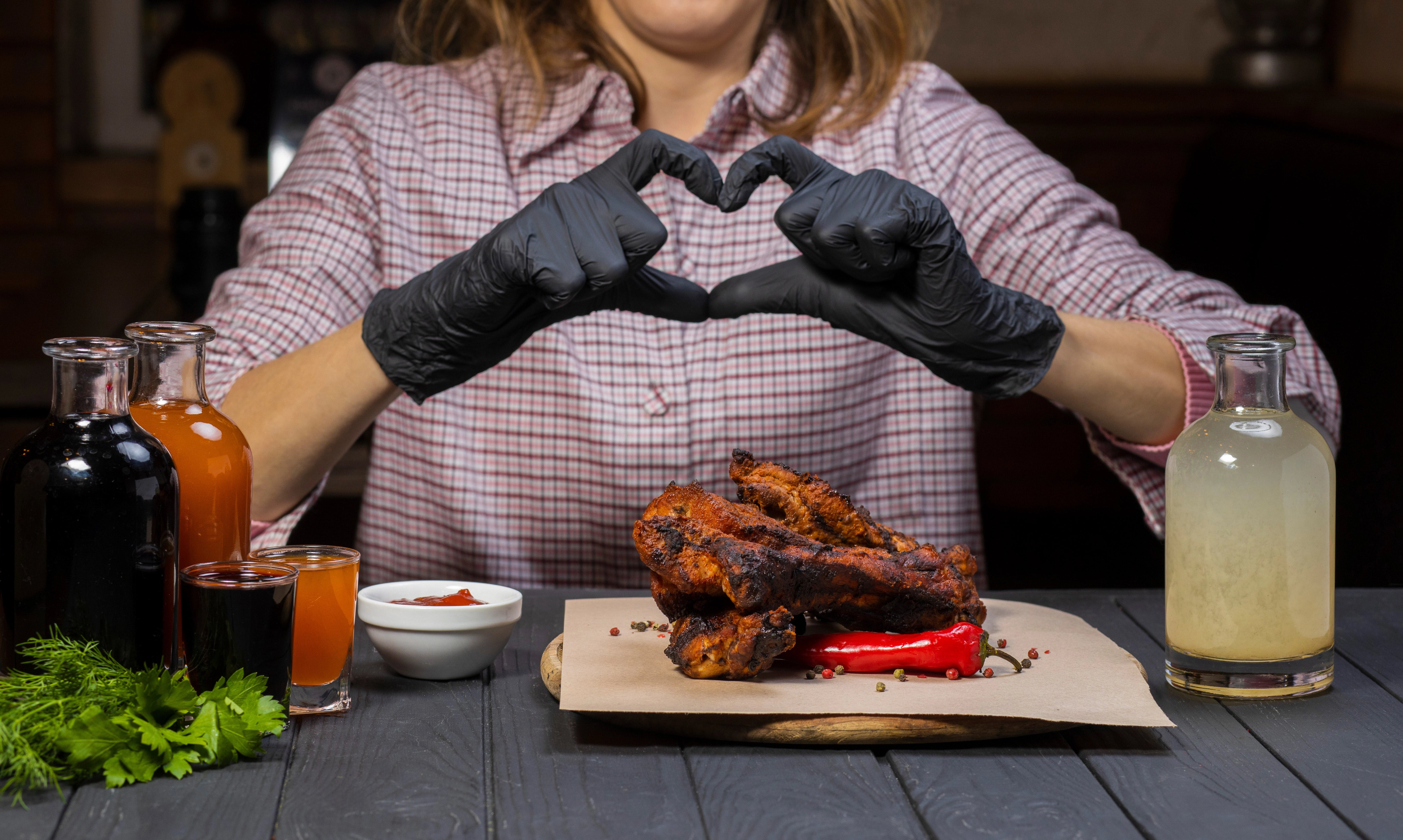 Frau formt mit schwarzen Handschuhen ein Herz, während Grillgut vor ihr liegt.