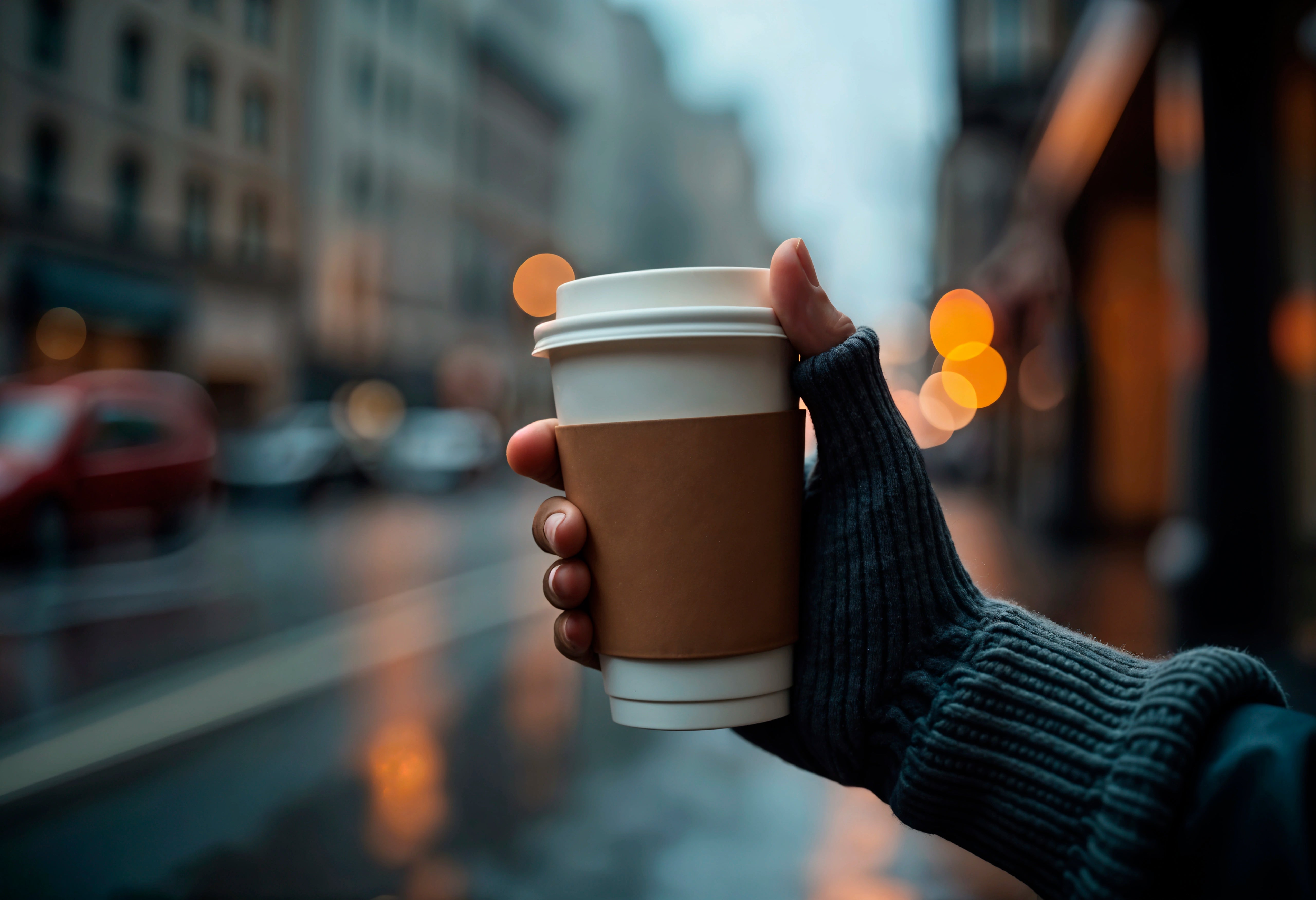 Hand, die einen Kaffeebecher to go vor einem unscharfen urbanen Hintergrund hält.